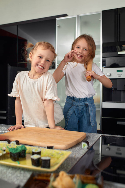 Δύο μικρά κορίτσια αδέλφια διασκεδάζουν και τρώνε στην κουζίνα στο σπίτι με γιαπωνέζικο φαγητό - Φωτογραφία, εικόνα