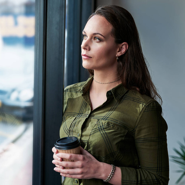 Νέες δυνατότητες προκύπτουν κάθε νέα ημέρα. Μια ελκυστική νεαρή επιχειρηματίας που πίνει καφέ και κοιτάζει έξω από το παράθυρο του γραφείου της. - Φωτογραφία, εικόνα