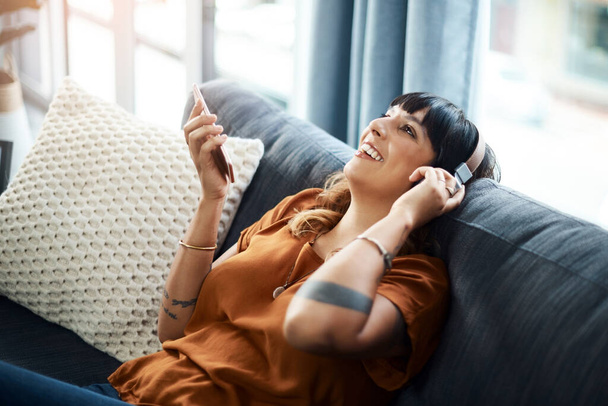 A música pode fazer você se sentir mais otimista e positivo sobre a vida. uma jovem mulher usando fones de ouvido enquanto usa seu celular em casa - Foto, Imagem