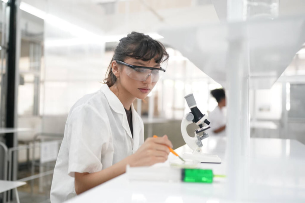Giovane dottoressa in uniforme che lavora con microscopio effettuando analisi presso l'ufficio di laboratorio - Foto, immagini