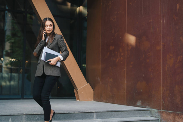 Confident beautiful caucasian businesswoman in a jacket standing holding a laptop talking on the phone with her client outside the office. Building, city - 写真・画像