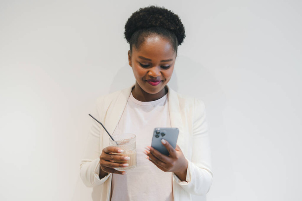 Photo of cheerful Afro American woman holding smartphone device, enjoying drinking beverage isolated on white wall background. People lifestyle. Social media - Fotoğraf, Görsel