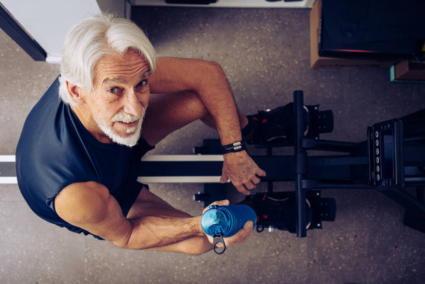 Indoor portrait of senior man working out on his rowing machine in his home office. - Fotó, kép