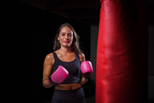 Pretty dark haired girl boxing a punching bag in a dark and moody setting. She is wearing boxing gloves and is fully focused on her training or exercise routine.  - Fotografie, Obrázek