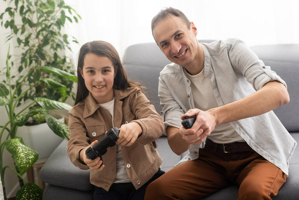 семейная, игровая и развлекательная концепция - счастливый отец и маленькая дочь с геймпадами, играющими дома в видеоигры. - Фото, изображение