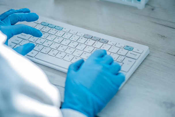 A female researcher sits at a workplace in a laboratory, behind a personal computer monitor. Against the background research statistics. Pharmaceutical medical worker in protection works at keyboard. - Foto, imagen