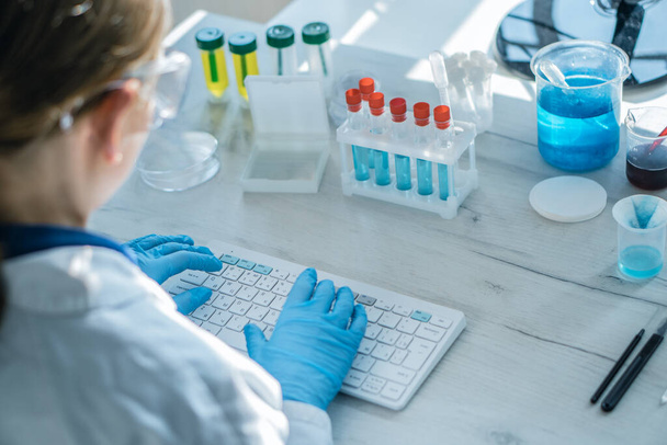 A female researcher sits at a workplace in a laboratory, behind a personal computer monitor. Against the background research statistics. Pharmaceutical medical worker in protection works at keyboard. - Photo, Image