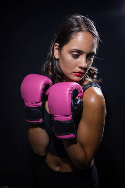 Pretty dark haired girl boxing a punching bag in a dark and moody setting. She is wearing boxing gloves and is fully focused on her training or exercise routine.  - Foto, Imagen