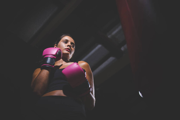 Pretty dark haired girl boxing a punching bag in a dark and moody setting. She is wearing boxing gloves and is fully focused on her training or exercise routine.  - Фото, зображення