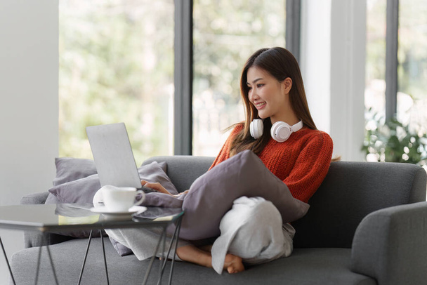 Beautiful Asian Woman smile and relaxing at home and using laptop computer sitting on cozy sofa. - Photo, image