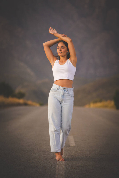 pretty young woman with dark hair on a deserted road. She is dressed in trendy and fashionable attire, including jeans and a stylish crop top. - Foto, afbeelding