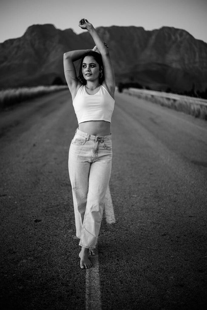 pretty young woman with dark hair on a deserted road. She is dressed in trendy and fashionable attire, including jeans and a stylish crop top. - Zdjęcie, obraz