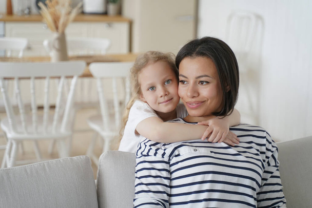 Blonde cute little girl hugs mom. Affectionate carefree family has happy moments together. Young spanish mother and european daughter on sofa in living room. Concept of child adoption. - Foto, afbeelding