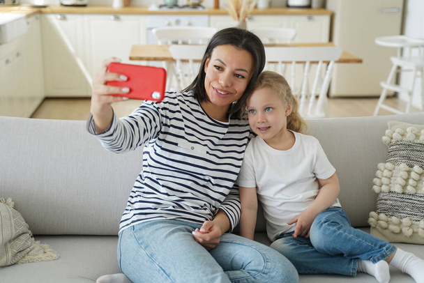 Mom and foster daughter chatting with father by video call, sitting on couch at home. Smiling mother holding phone, taking selfie with little child for social network together. Adoption and motherhood - Valokuva, kuva