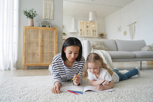Mom and her kid are drawing together. Young caucasian woman and little daughter lying on floor together and coloring with markers. Happy family has good time at home. Childhood and parenthood concept. - Foto, Imagen