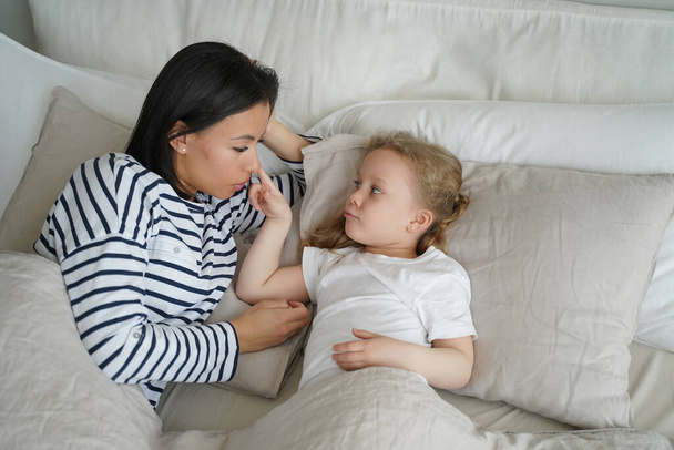 Mother lying in comfortable bed with small foster girl in bedroom together. Mom wake up little daughter after sleeping in the morning at home. Healthy sleep in children, childcare. - Valokuva, kuva