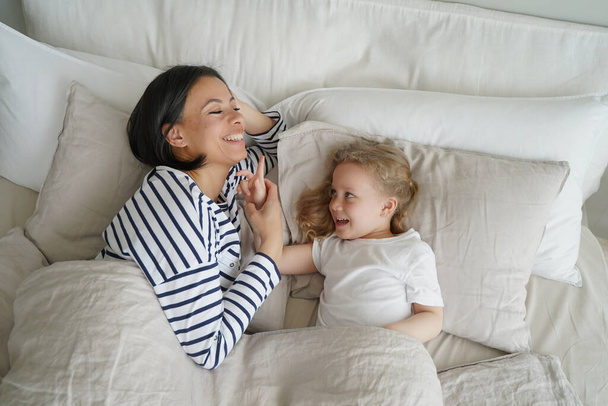 幼い母親は寝室で娘と一緒に休んでいる。白人の女性と子供は一緒に笑顔で眠りに落ちています。母親と子供が目覚めつつある。子育てと関係の概念. - 写真・画像