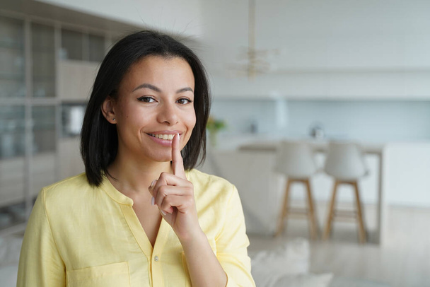 Smiling female puts finger on lips, makes hush gesture at home. Happy woman share secret private information, ask silence, looking at camera. Unique sale offer, big discounts in online store. - Фото, изображение