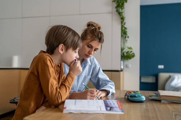 Young mother teaching teenage boy at home, mom helping puzzled kid son with difficult school task, supporting child in remote learning. Woman freelancer balancing parenting and remote work. Homeschool - Photo, Image