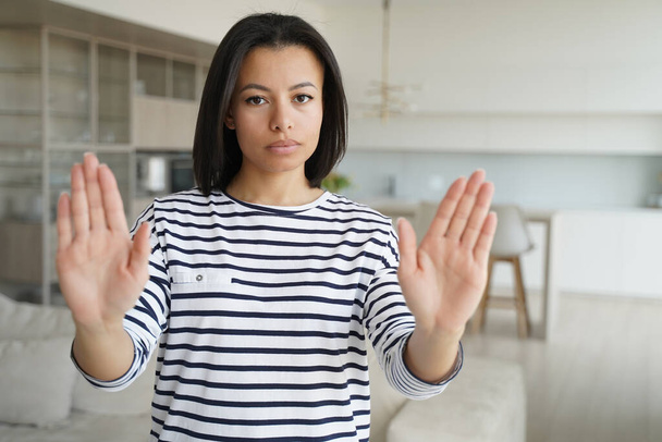 Young woman showing stop gesture by palms, warning sign of prohibition or rejection, looking at camera at home. Serious strict female protesting against domestic violence, abuse, gender discrimination - Foto, afbeelding