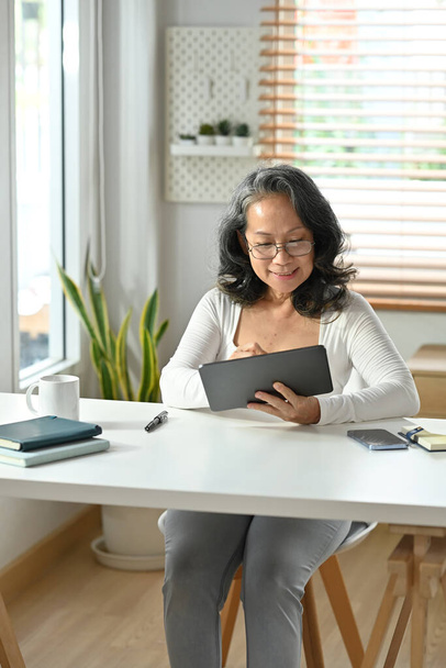 Portret zrelaksowanej dojrzałej kobiety korzystającej z aplikacji mobilnych, przeglądającej media społecznościowe siedząc w biurze. - Zdjęcie, obraz