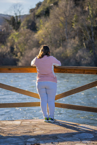 zdjęcie pięknej kobiety w średnim wieku w różowym swetrze i jasnoniebieskich spodniach na brzegu rzeki o zachodzie słońca - Zdjęcie, obraz