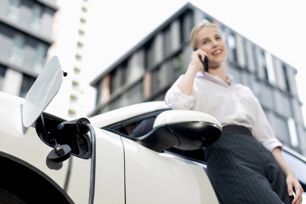 Focus charging-electric car with EV charger at charging station with blur businesswoman talking on phone with residential building in background as progressive lifestyle concept. - 写真・画像
