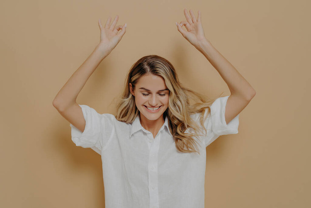 Carefree lovely female in white shirt with blonde hair enjoying nice day, keeping eyes closed and raising arms, making okay gesture with both hands and demonstrating positive emotions - Photo, Image