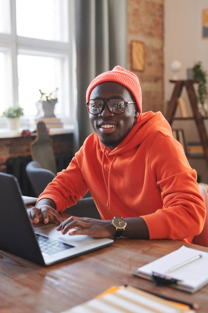 Vertical portrait of joyful African American guy wearing bright orange outfit sitting at desk at home surfing Internet on laptop smiling at camera - Fotografie, Obrázek