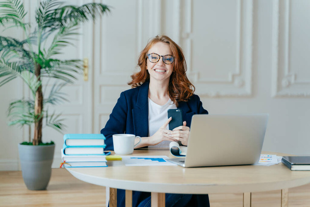 Horizontal shot of optimistic woman installs application on modern cell phone, checks notification, surrounded with paper documents, works on laptop computer, has red hair, dressed formally. - Foto, afbeelding