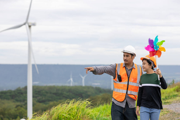 Engineer with his son holding windmill toy on a wind farm atop a hill or mountain. Progressive ideal for the future production of renewable, sustainable energy. Energy generated from wind turbine. - Foto, Bild