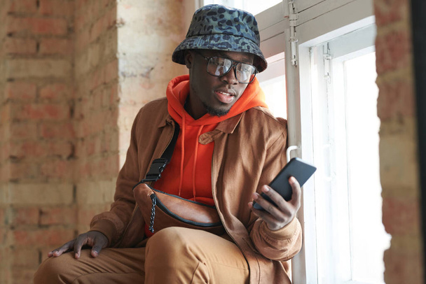 Horiozntal medium portrait of handsome young Black man wearing casual outfit and eyeglasses sitting on window sill having video call on smartphone - Фото, изображение