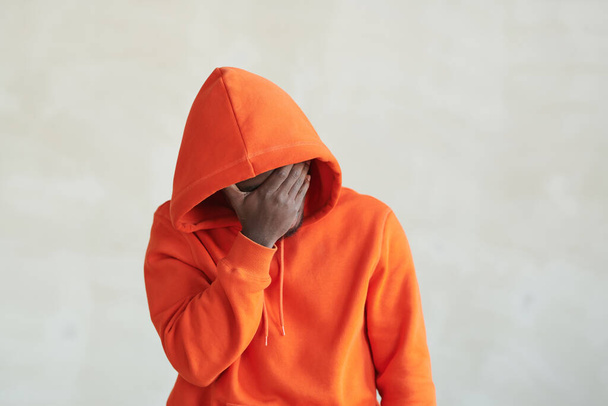 Horizontal medium studio conceptual portrait of unrecognizable African American man wearing orange hoodie covering his face with hand, copy space - Фото, зображення