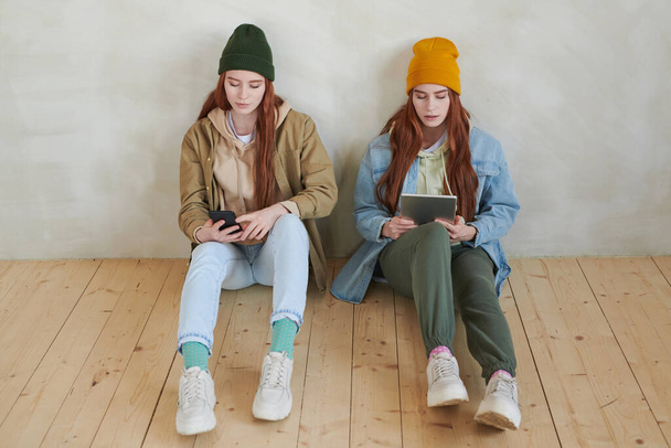 High angle view studio portrait of young twin sisters wearing fashionable casual clothes sitting on wooden floor using gadgets - Zdjęcie, obraz