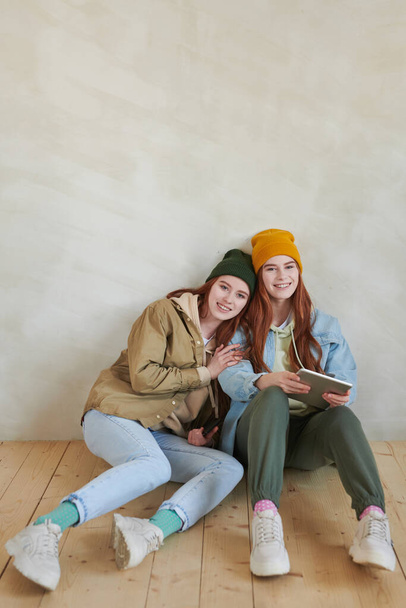 Vertical high angle studio portrait of joyful young female twins wearing stylish casual outfits sitting together on flloor smiling at camera - Foto, imagen