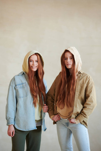 Vertical medium long portrait of two beautiful young Caucasian female twins wearing denim jackets, jeans and hoodies looking at camera - Zdjęcie, obraz