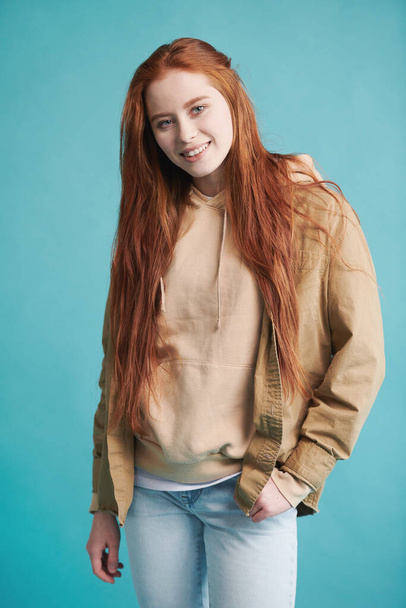 Vertical medium studio portrait of joyful young woman with long red hair wearing casual clothes posing on camera against blue wall background - Photo, Image