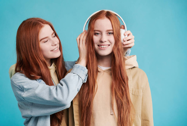 Waist up portrait of lovely red-haired Cacuasian woman putting white headphones on her twin sisters head, blue background - Фото, зображення