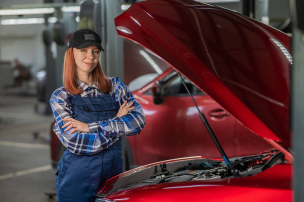 A woman auto mechanic stands at the open hood of a car with her arms crossed over her chest - Фото, зображення