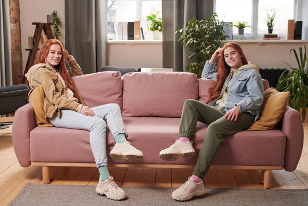 Wide shot of two beautiful young Caucasian twin sisters with long red hair wearing casual outfits sitting relaxed on sofa in living room - Zdjęcie, obraz