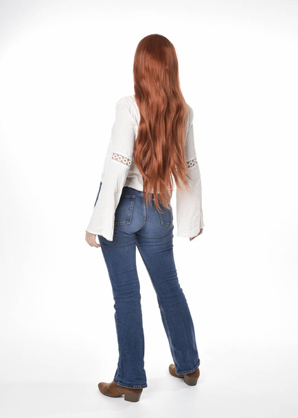 full length portrait of beautiful woman model with long red hair, wearing casual outfit white blouse  top and denim jeans, isolated on white studio background. Backwards standing pose, walking away from camera. - Photo, Image