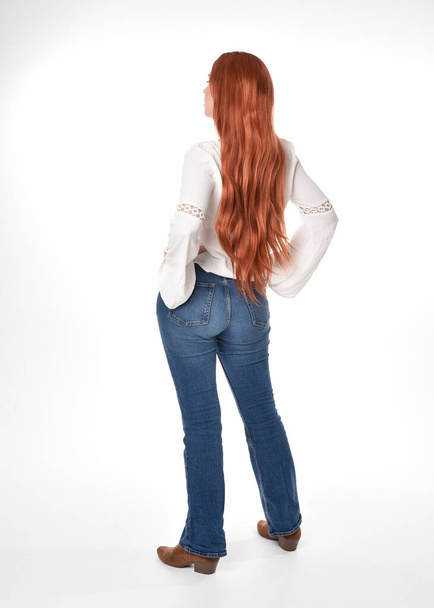 full length portrait of beautiful woman model with long red hair, wearing casual outfit white blouse  top and denim jeans, isolated on white studio background. Backwards standing pose, walking away from camera. - Foto, Imagen