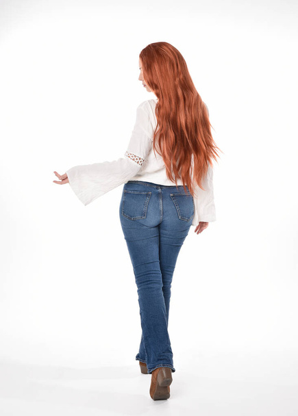Ganzkörperporträt der schönen Frau Modell mit langen roten Haaren, trägt legeres Outfit weiße Bluse Top und Jeans, isoliert auf weißem Studiohintergrund. Rückwärts stehend posieren, von der Kamera weggehen. - Foto, Bild