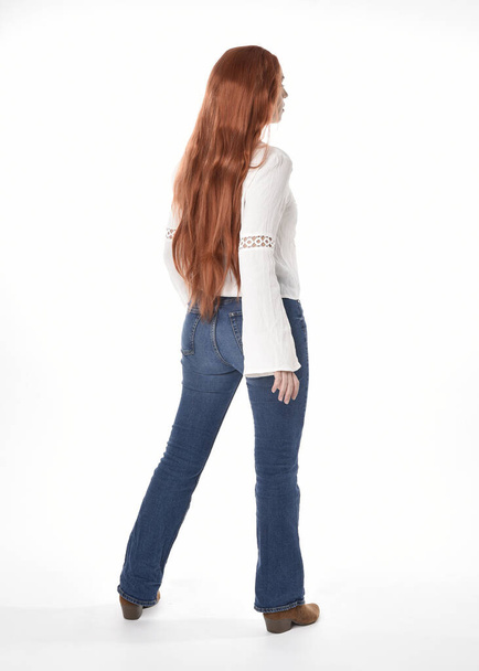 полноразмерный портрет красивой женщины-модели с длинными рыжими волосами, в повседневной одежде, белой блузке и джинсах в джинсах, выделенных на белом фоне студии. Стоя спиной, отойдя от камеры. - Фото, изображение