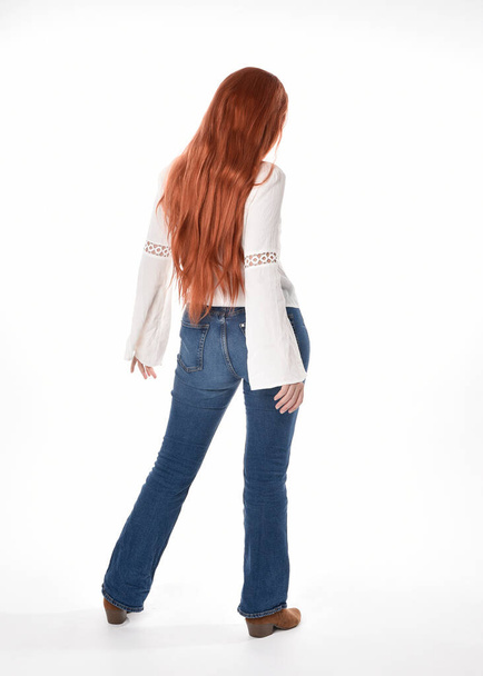 портрет красивої жінки-моделі з довгим рудим волоссям, в повсякденному вбранні біла блузка зверху і джинсові джинси, ізольовані на білому студійному фоні. Позиція позаду, що стоїть, відійшовши від камери
. - Фото, зображення