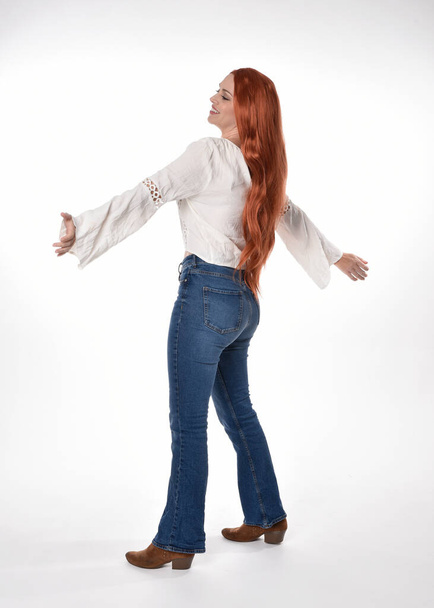 pełnowymiarowy portret pięknej kobiety modelki z długimi czerwonymi włosami, ubranej w luźny strój biała bluzka i jeansy jeansowe, odizolowanej na białym tle pracowni. Stojąca do tyłu poza, odchodząca od kamery. - Zdjęcie, obraz