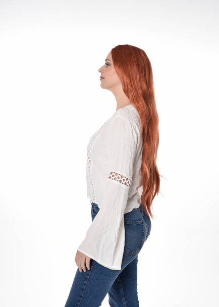  Κοντινό πλάνο πορτρέτο της όμορφης γυναίκας μοντέλο με μακριά κόκκινα μαλλιά, φορώντας casual σύνολο λευκό μπλούζα κορυφή και τζιν τζιν τζιν. Τοποθέτηση με χειρονομίες βραχίονα, απομονωμένη σε λευκό φόντο στούντιο.  - Φωτογραφία, εικόνα