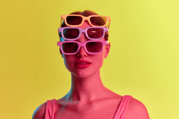 Moda. Portret młodej dziewczyny noszącej kilka okularów przeciwsłonecznych i patrzącej przez nie na kamerę ponad żółto-pomarańczowym tłem w neonowym świetle. Pojęcie piękna, młodości, ludzkich emocji, nastroju, reklamy - Zdjęcie, obraz