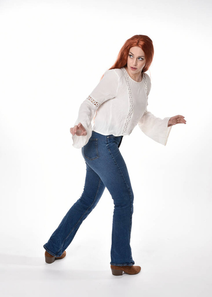full length portrait of beautiful woman model with long red hair, wearing casual outfit white blouse  top and denim jeans, isolated on white studio background. standing pose, walking towards the camera with gestural hand poses. - Fotoğraf, Görsel