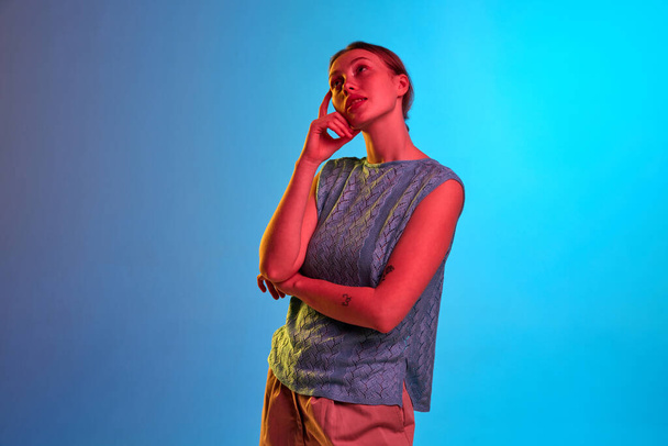 Dreamy, thoughtful, calm. Shot of pensive girl with holded hands standing and thinking about something over blue background in neon light. Concept of beauty, human emotions, mood, ad - Foto, immagini